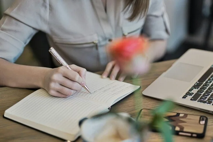 Mujer escribiendo en una libreta junto a un portátil y una rosa roja